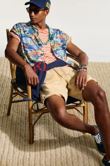 Man in casual summer fashion sits in a bamboo chair wearing a floral shirt, shorts, baseball cap, sunglasses, necklace and bracelets. 