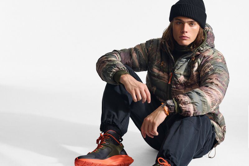 A person wearing a camouflage puffer jacket and black beanie sits on the floor with orange and black high-top sneakers in a studio shot.