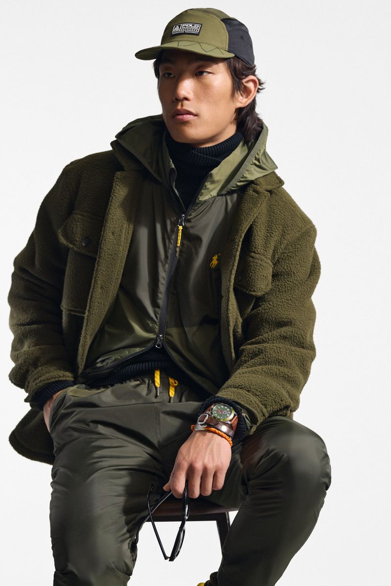 An Asian male model poses on a stool, showcasing an olive-green sherpa jacket, zip-up vest, matching trousers, and cap against a plain backdrop.