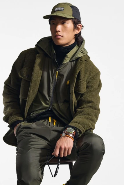 An Asian male model poses on a stool, showcasing an olive-green sherpa jacket, zip-up vest, matching trousers, and cap against a plain backdrop.