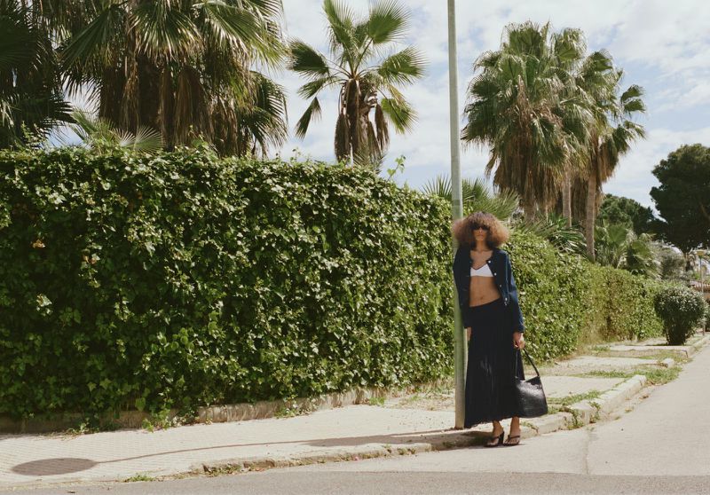 A woman with afro-style hair poses in a bra top, jacket, and skirt next to a green hedge, with palm trees in the background on a sunny day.