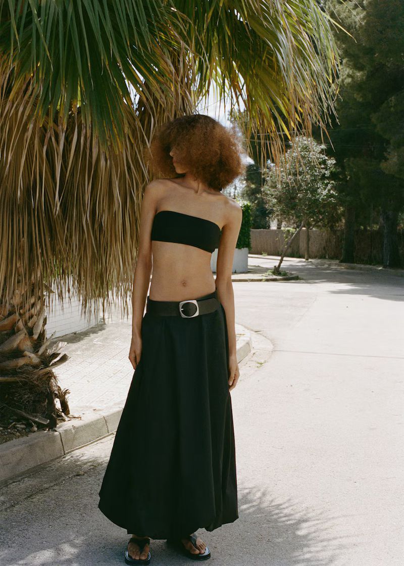 Woman with afro wears black bandeau top and long skirt outside near palm tree in natural light, fashion editorial, waist-up shot.