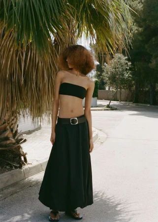 Woman with afro wears black bandeau top and long skirt outside near palm tree in natural light, fashion editorial, waist-up shot.