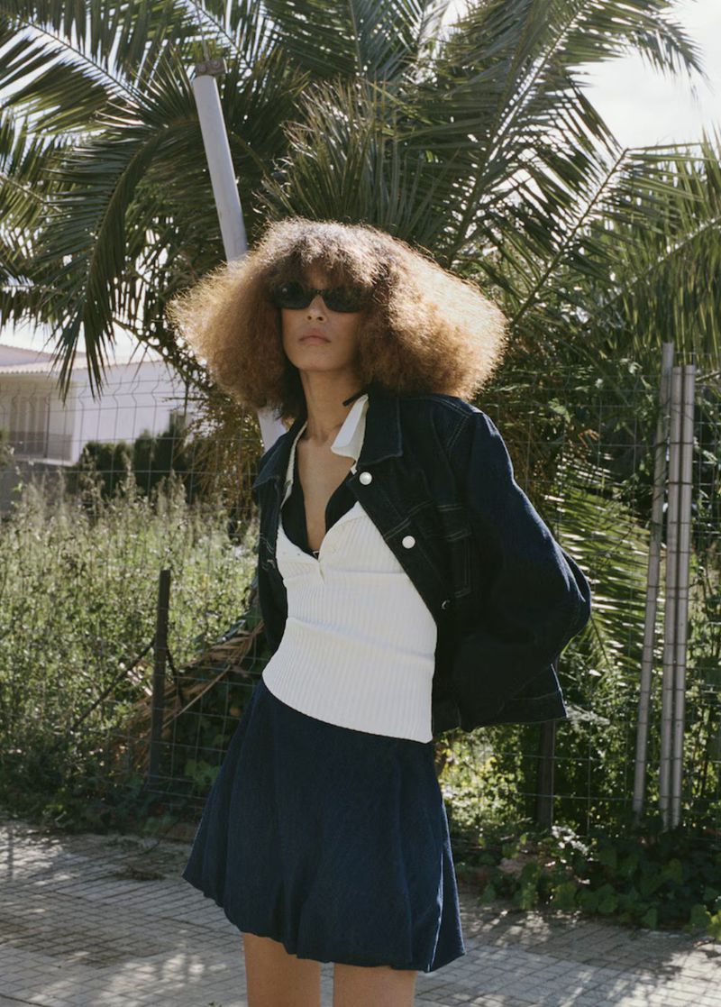 Woman with curly hair poses in a dark jacket, white top, and dark skirt on a sidewalk with palm trees in the background.