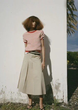 Fashion shot of a female model in a red-and-white striped top and khaki skirt, posing against a white wall with greenery in the background.