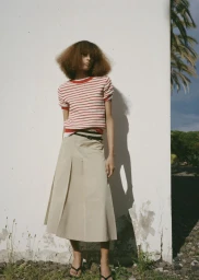 Model wearing a red and white striped knit top, beige pleated midi skirt, and black sandals against an outdoor white wall.
