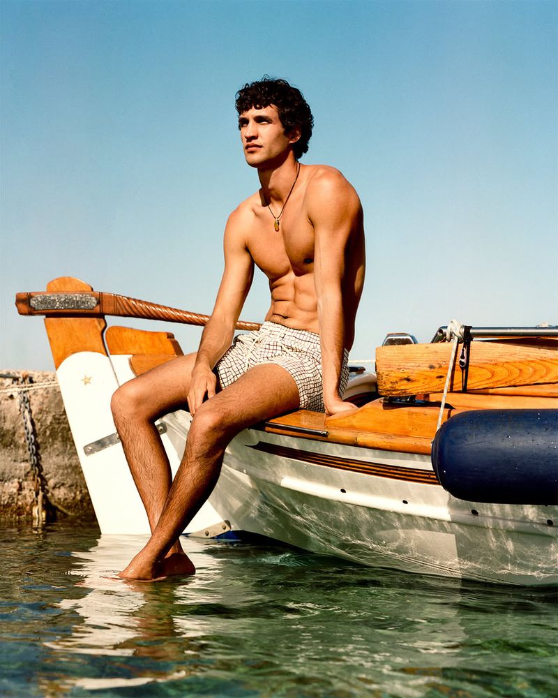 Male model sits shirtless on a wooden boat in clear water, looking to the side under a bright blue sky on a sunny day.