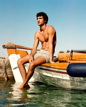 Male model sits shirtless on a wooden boat in clear water, looking to the side under a bright blue sky on a sunny day.