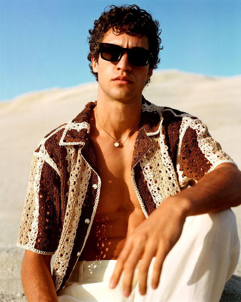 Young man poses in a crochet shirt and white pants against a sandy backdrop, wearing sunglasses and a necklace under a blue sky.