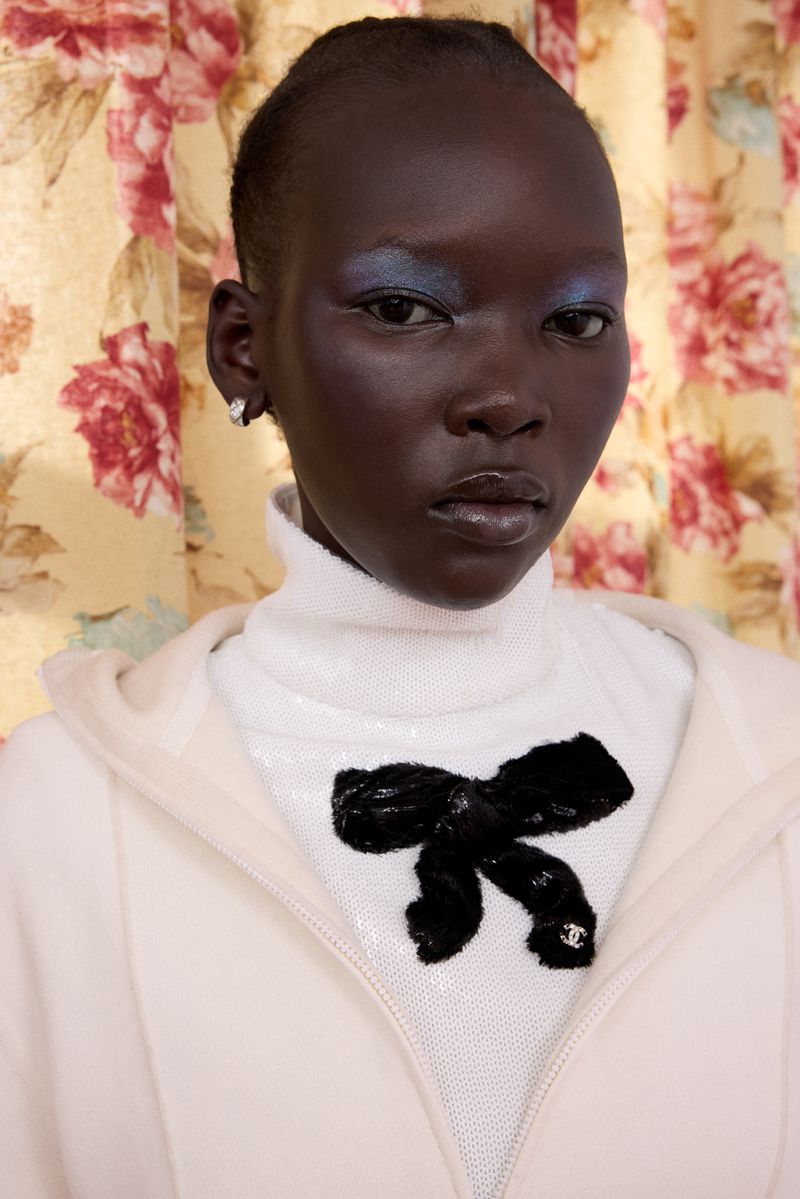 Close-up of a dark-skinned woman with blue eyeshadow wearing a white sequined top with a black bow, against a floral background.
