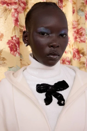 Close-up of a dark-skinned woman with blue eyeshadow wearing a white sequined top with a black bow, against a floral background.