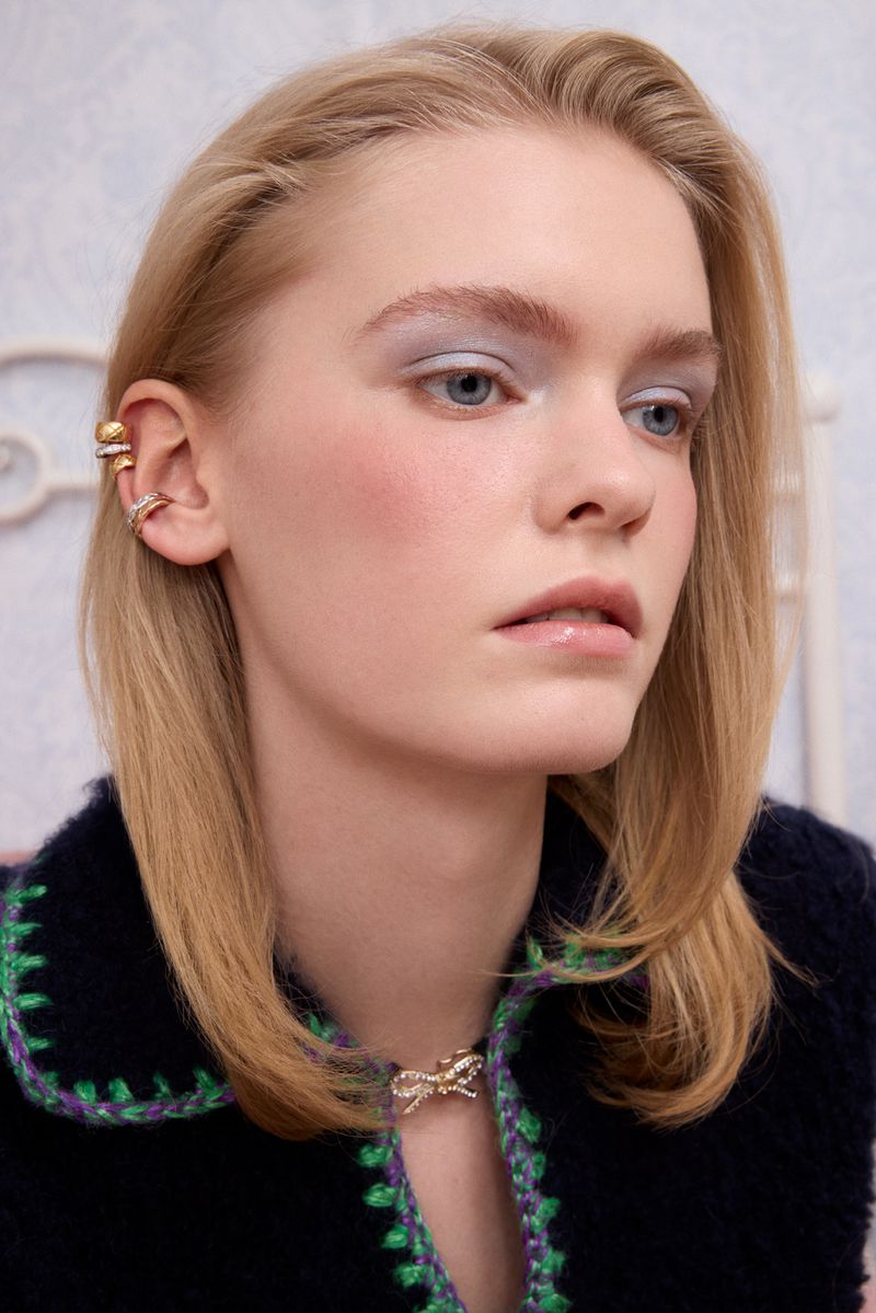 Young woman with blonde hair wearing gold jewelry and a dark blue sweater with green trim poses indoors in a portrait style shot.