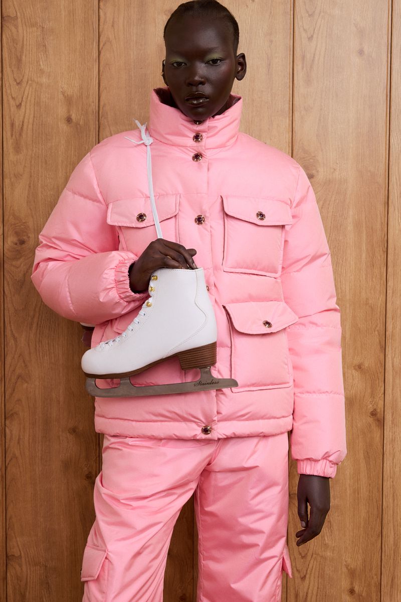Woman in pink puffer suit holding ice skates against a wood paneled wall in a studio-lit setting. Fashion and sportswear editorial.