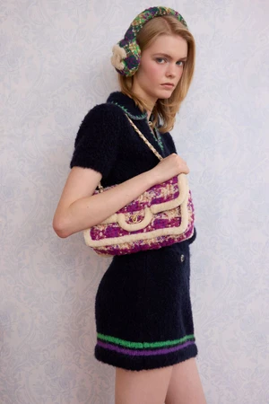 Model in a navy knit dress and tweed earmuffs holding a purple and cream tweed shoulder bag against patterned wallpaper.