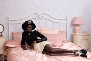 Model reclining across a bed dressed in pink satin bedding wearing a black Chanel trapper hat, black sweater, and cream skirt.