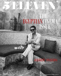 Egyptian actor Asser Yassin sits on a curved cushioned bench wearing sunglasses, a light collared shirt, trousers, and dark slip-ons.