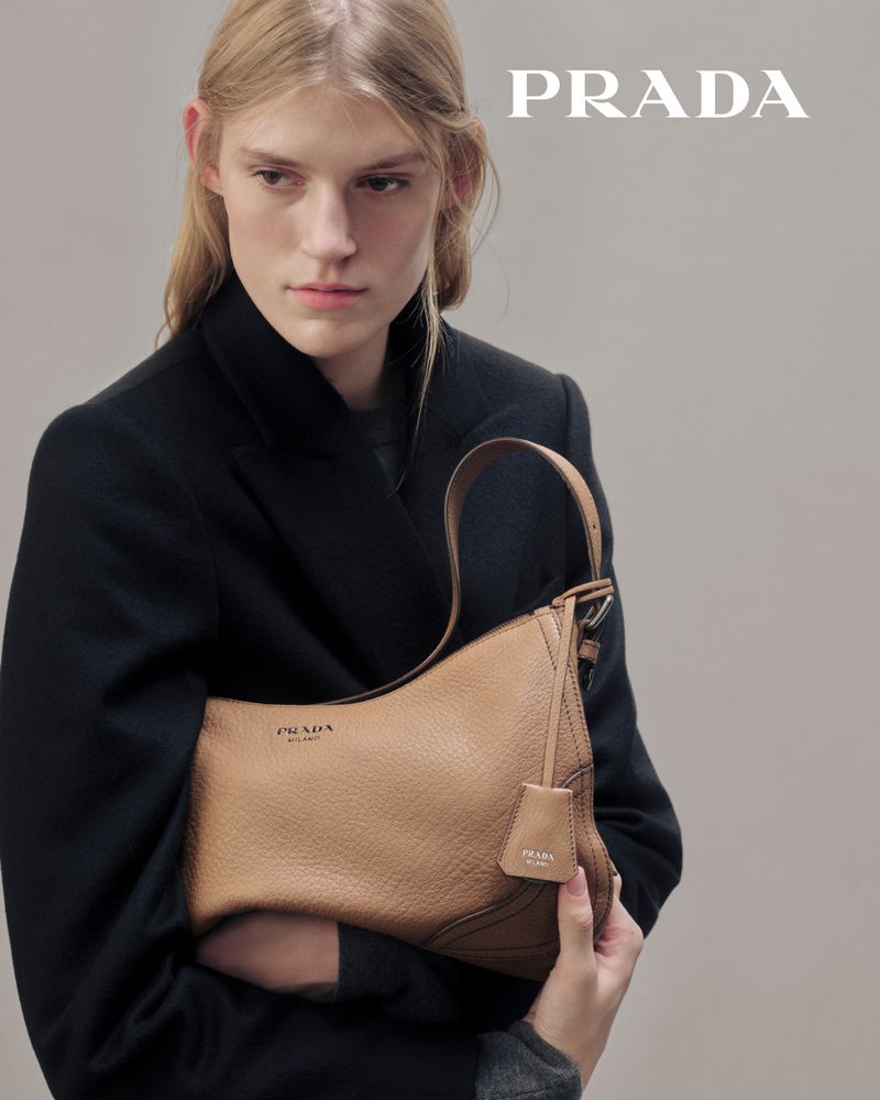 Fashion shot of a fair-skinned female model showcasing a tan leather Prada handbag against a light grey backdrop with the brand logo visible.
