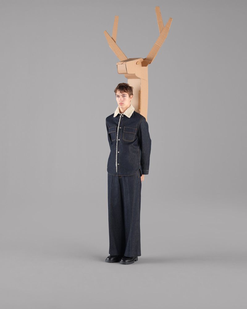 A young adult poses in a dark denim outfit next to a cardboard deer head and antlers against a neutral gray backdrop in a studio.