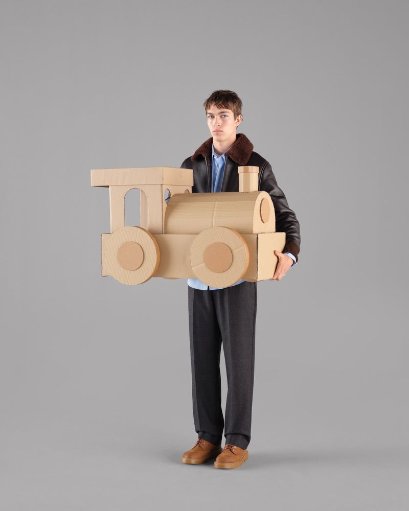Young adult man holding a large cardboard toy train in a studio environment, wearing a leather jacket and dress pants.