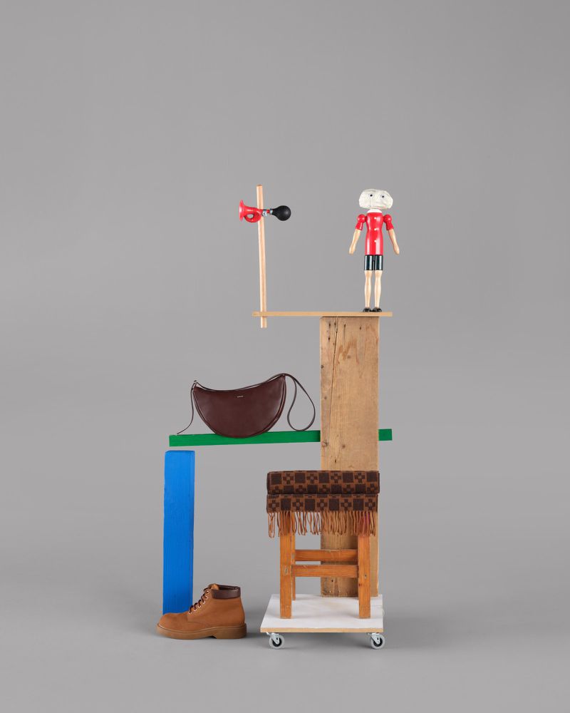 A still life features a brown leather shoulder bag, brown leather boot, a toy doll, and a child's stool against a gray background.
