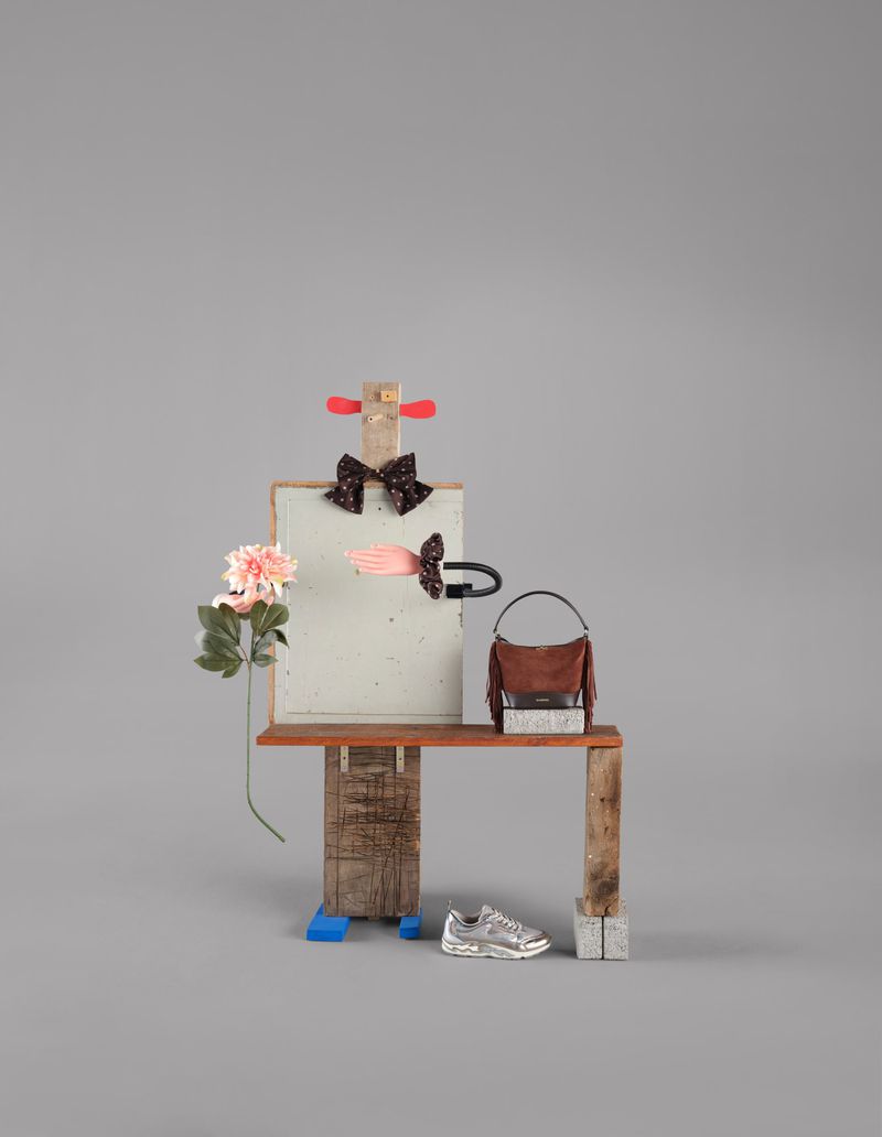 Still life composition of a handbag, sneakers, and floral element arranged on a wooden structure against a grey backdrop.