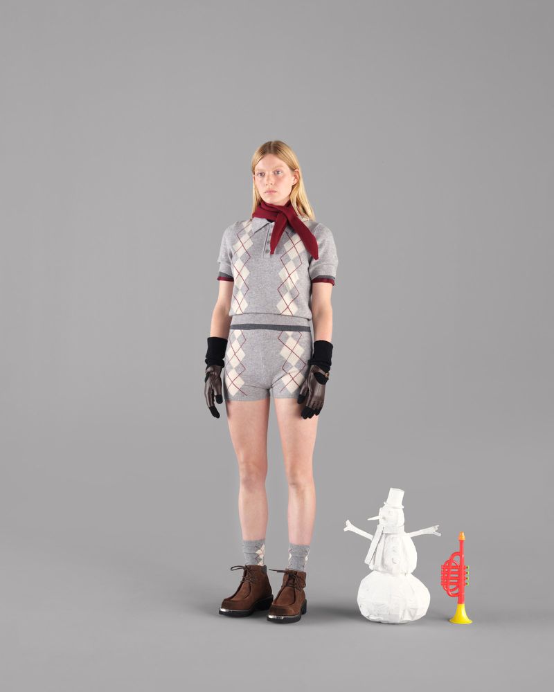 Blonde woman in argyle polo and shorts, gloves, boots, scarf poses with snowman and toy trumpet on gray studio backdrop.
