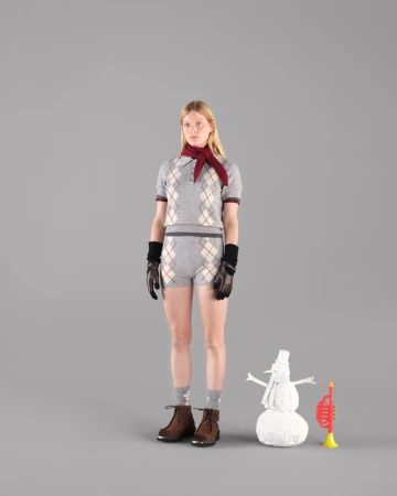 Blonde woman in argyle polo and shorts, gloves, boots, scarf poses with snowman and toy trumpet on gray studio backdrop.