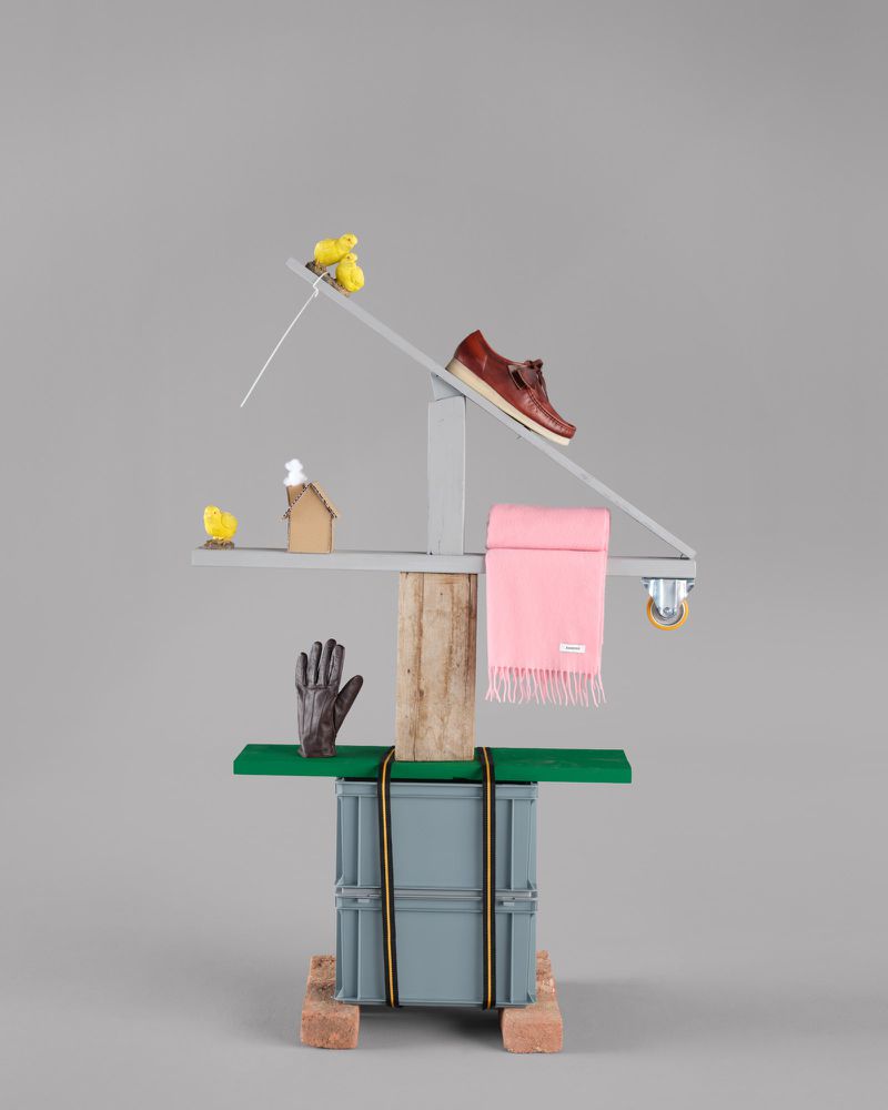 A still life photograph features a brown shoe atop a whimsical, balanced structure of planks, box, and wooden block on a grey background.