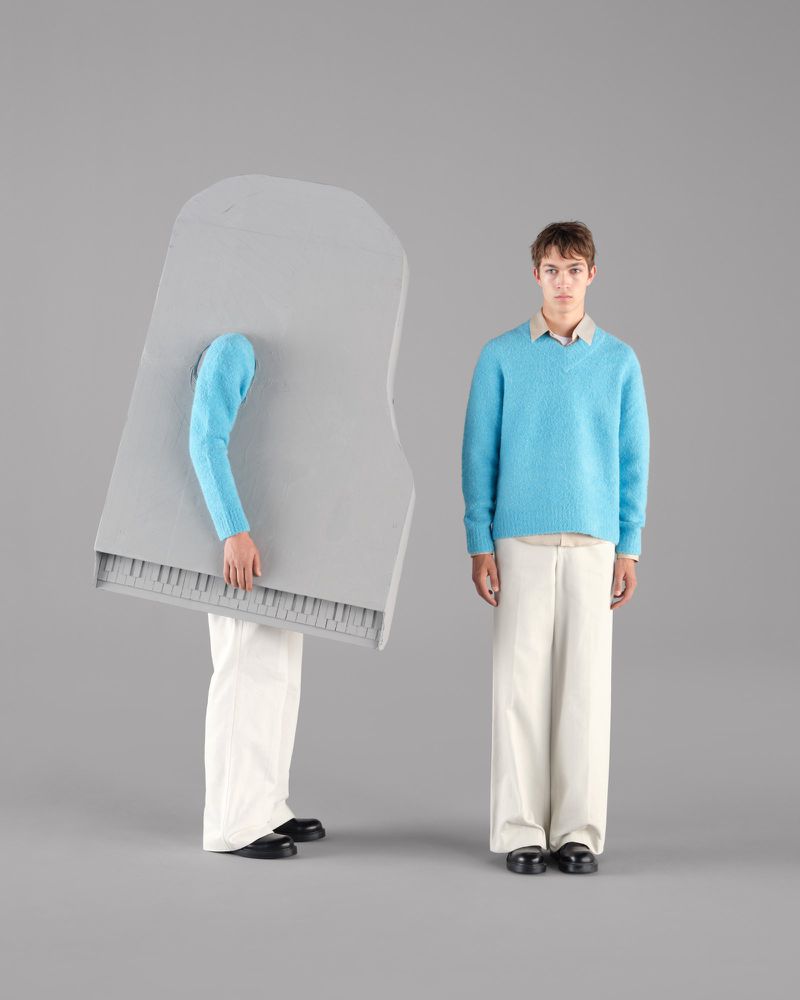 Two young men in studio setting, one dressed as a piano, wearing matching blue sweaters and beige trousers. Fashion and music concept.