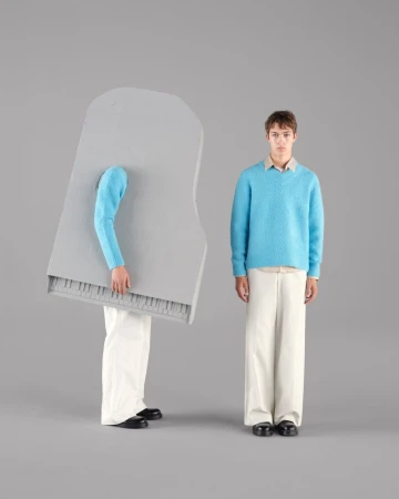 Two young men in studio setting, one dressed as a piano, wearing matching blue sweaters and beige trousers. Fashion and music concept.