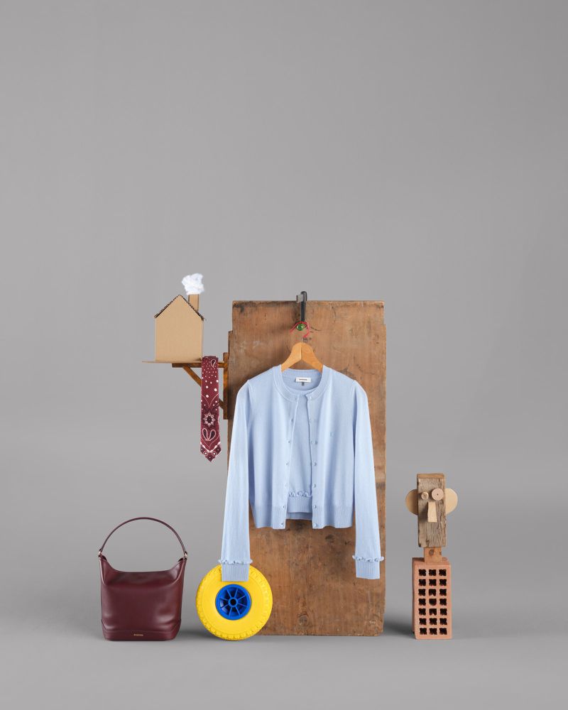 Studio shot of light blue cardigan, burgundy purse, and various props including a toy house and a yellow wheel on a grey background.