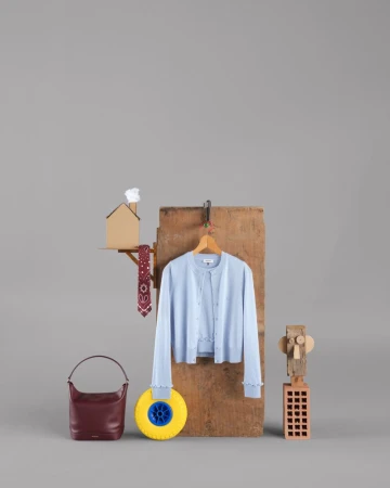 Studio shot of light blue cardigan, burgundy purse, and various props including a toy house and a yellow wheel on a grey background.