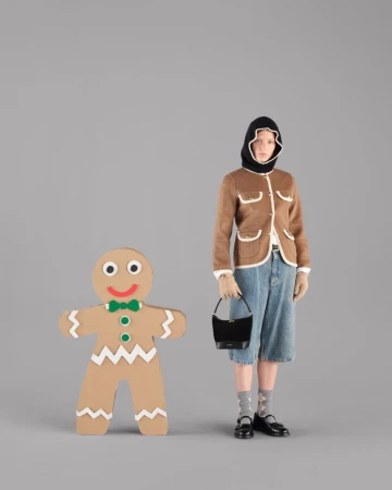 A woman in a brown jacket and denim shorts stands next to a large gingerbread man cutout against a gray background in a studio shot.