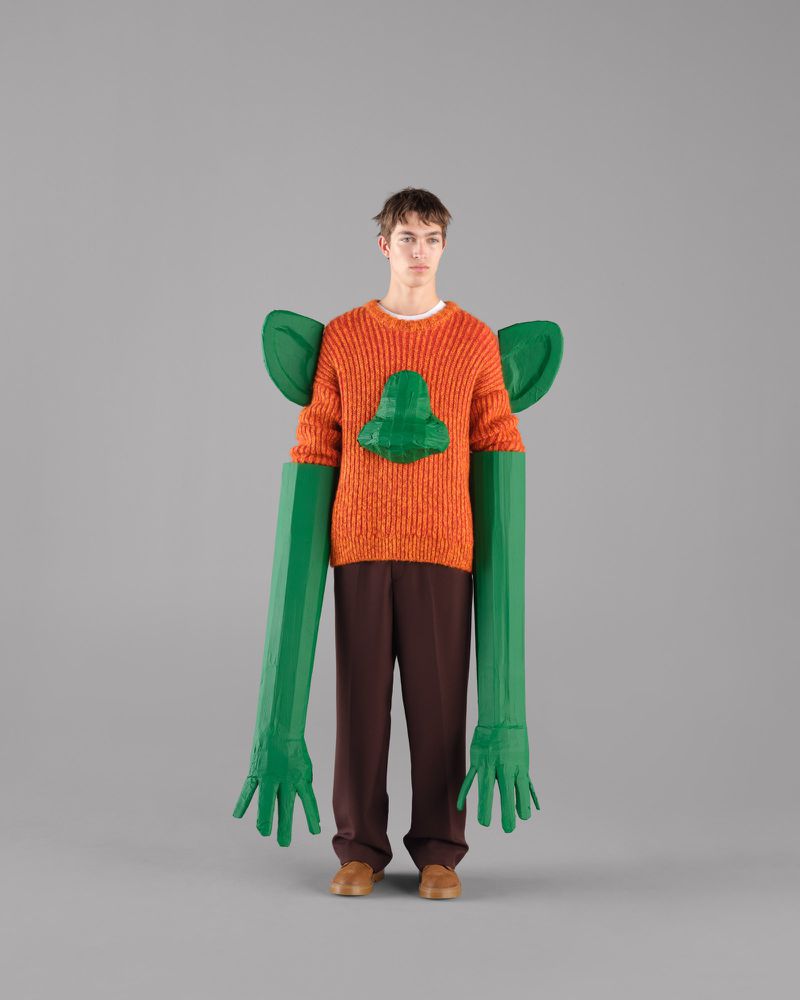 Male model in an orange knit sweater with 3D green accents poses against a grey backdrop for a fashion editorial shot.