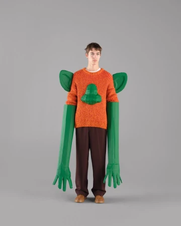 Male model in an orange knit sweater with 3D green accents poses against a grey backdrop for a fashion editorial shot.