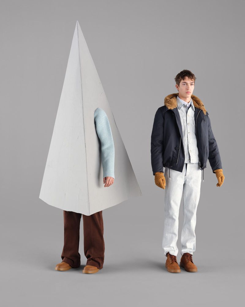 Two men pose against a grey background; one wears a navy bomber jacket, while the other stands inside a large geometric prop.