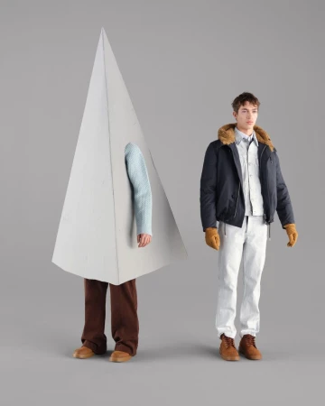Two men pose against a grey background; one wears a navy bomber jacket, while the other stands inside a large geometric prop.