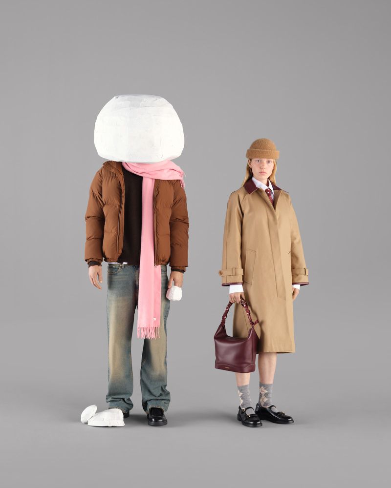 Two people wearing coats, one with a paper head and scarf, the other with a handbag, stand against a grey studio backdrop.