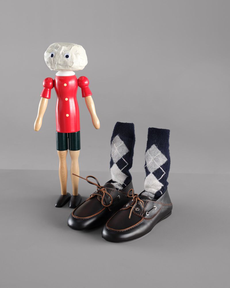 Wooden doll in red shirt standing next to pair of brown leather shoes with navy blue socks with argyle pattern on grey background.
