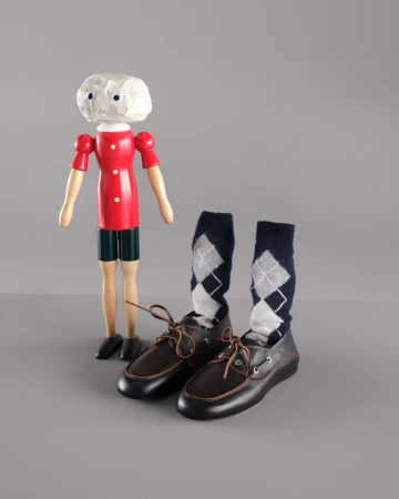 Wooden doll in red shirt standing next to pair of brown leather shoes with navy blue socks with argyle pattern on grey background.