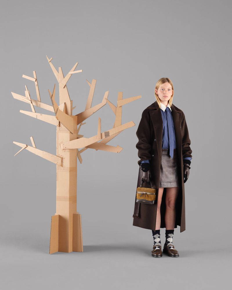A woman poses next to a cardboard tree in a studio, wearing a brown coat, blue blouse, grey skirt, patterned socks, and holding a purse.