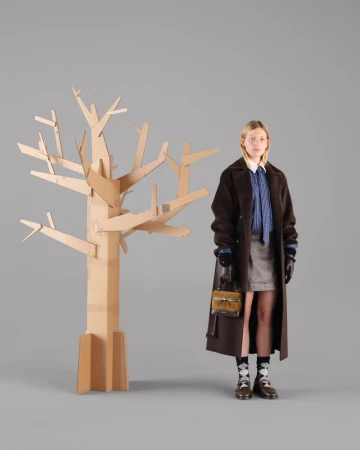 A woman poses next to a cardboard tree in a studio, wearing a brown coat, blue blouse, grey skirt, patterned socks, and holding a purse.