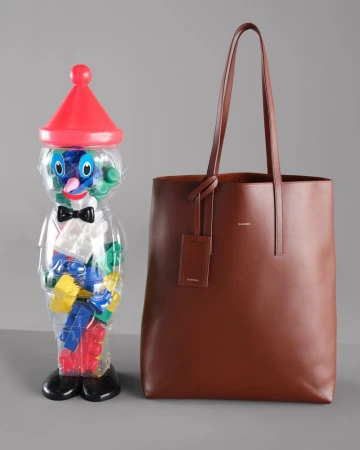 Brown leather tote bag with a plastic toy figure filled with blocks on a grey surface, studio shot.