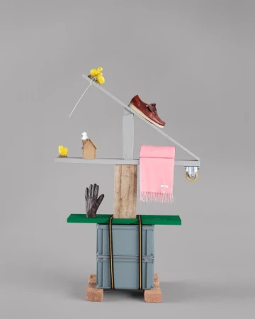Sculptural still life construction displaying a brown leather loafer, pink fringed scarf, and brown glove against a grey backdrop.