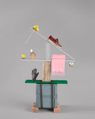 Sculptural still life construction displaying a brown leather loafer, pink fringed scarf, and brown glove against a grey backdrop.