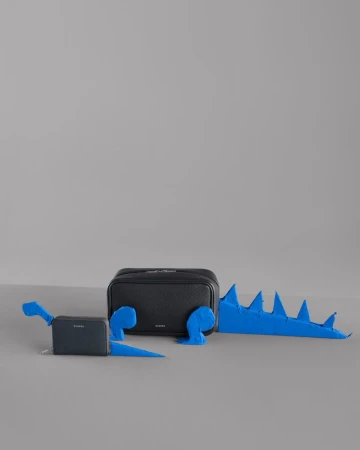 Black leather bag and small wallet styled with blue paper cutouts to resemble dinosaurs against a grey seamless studio background.