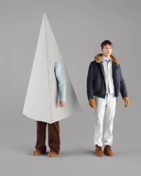 Model standing next to a figure encased in a large grey pyramid prop wearing a light blue sweater and brown trousers against a grey seamless background