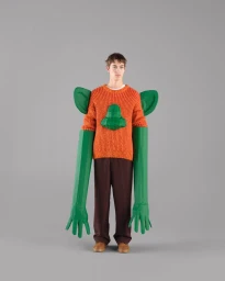 Model wearing an orange knit sweater with oversized sculptural green monster arms and brown trousers against a grey seamless background.
