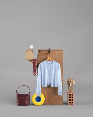 Light blue cardigan on a wooden block beside a burgundy handbag, yellow wheel, and cardboard house against a grey background.