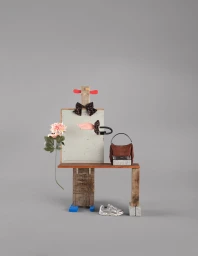 Anthropomorphic assemblage sculpture featuring a brown handbag, silver sneaker, pink flower, and bow tie against a grey studio backdrop.